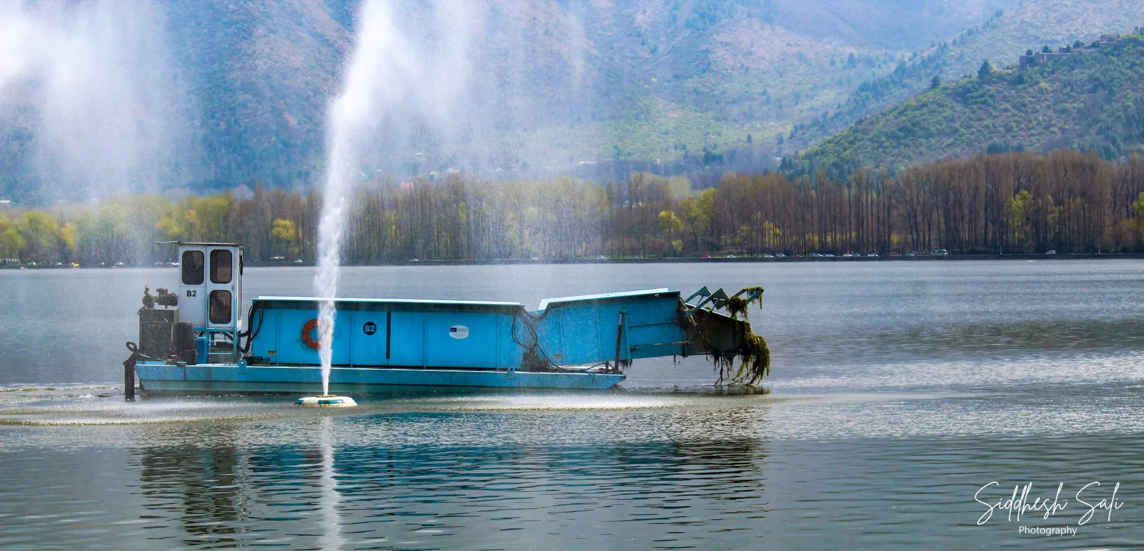 A blue barge on a lake throwing a tall jet of water, bare-branched trees along the far shore.