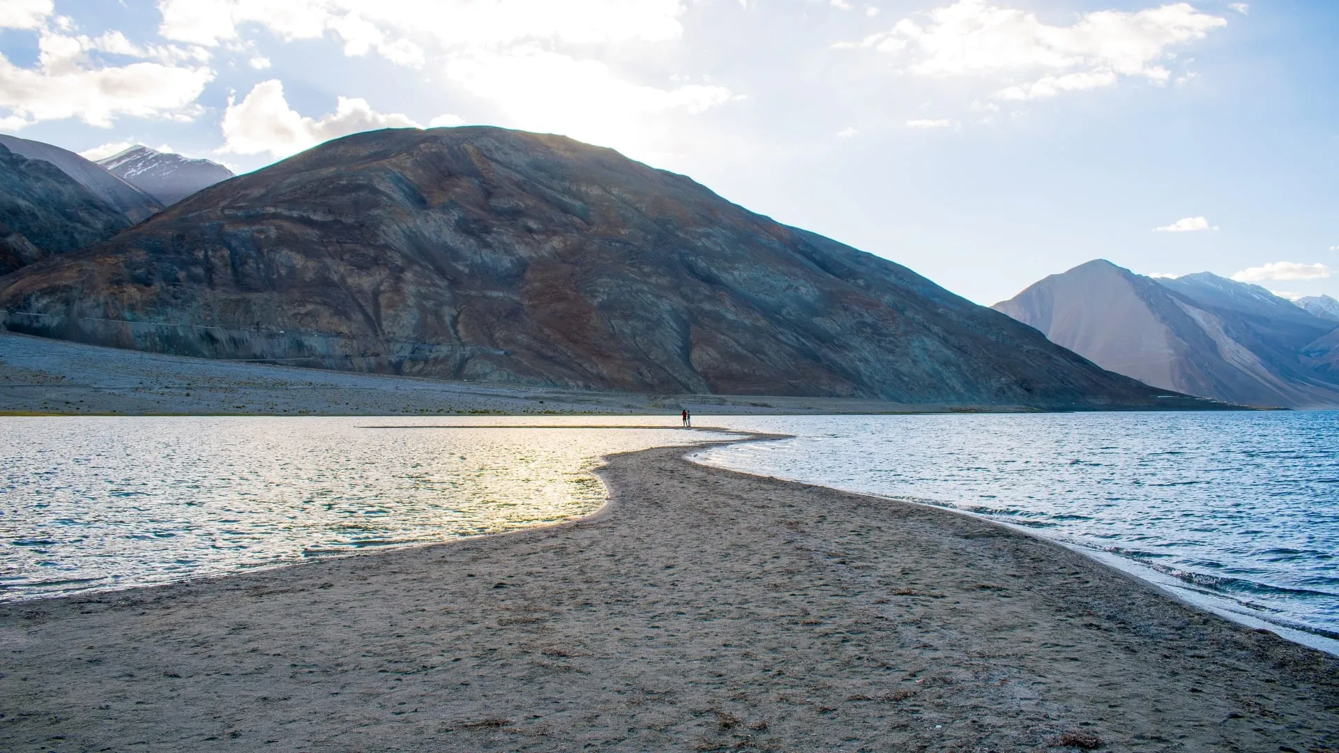 A narrow spit of pale sand curving out into flat water, a lone figure at its end.