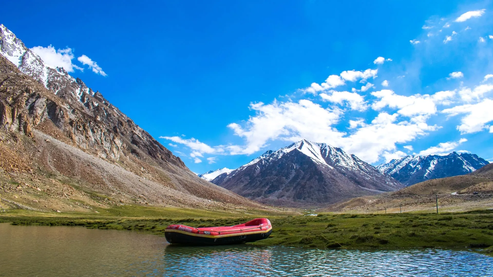 A small red boat moored on a green shore below snow-capped peaks and heavy cumulus.