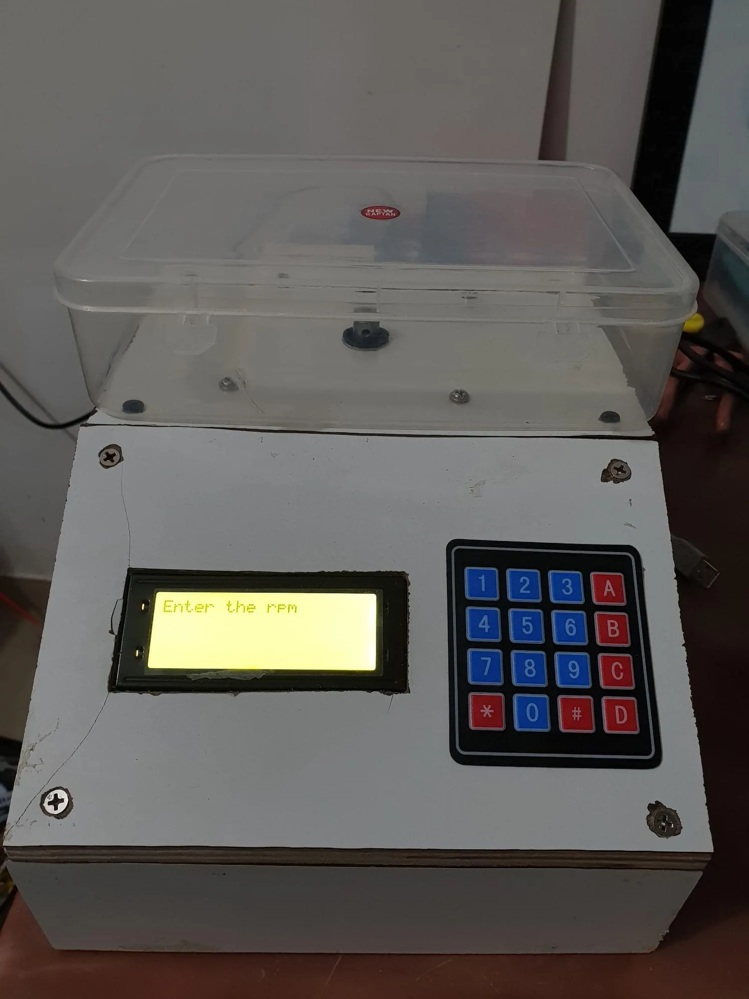 The finished spin coater: a pale metal enclosure with a backlit LCD and a red-and-blue keypad, a clear plastic chamber sitting on top.