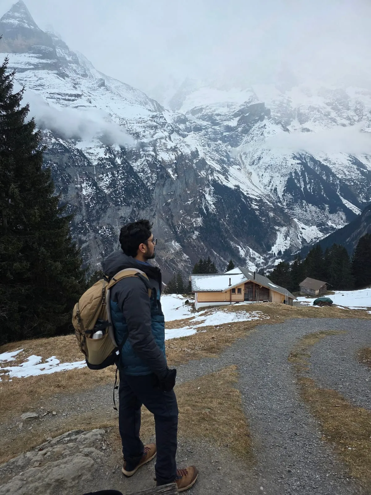 Siddhesh on a gravel path at Jungfraujoch, Switzerland, looking towards a wooden chalet below steep, snow-covered, cloud-hung peaks.
