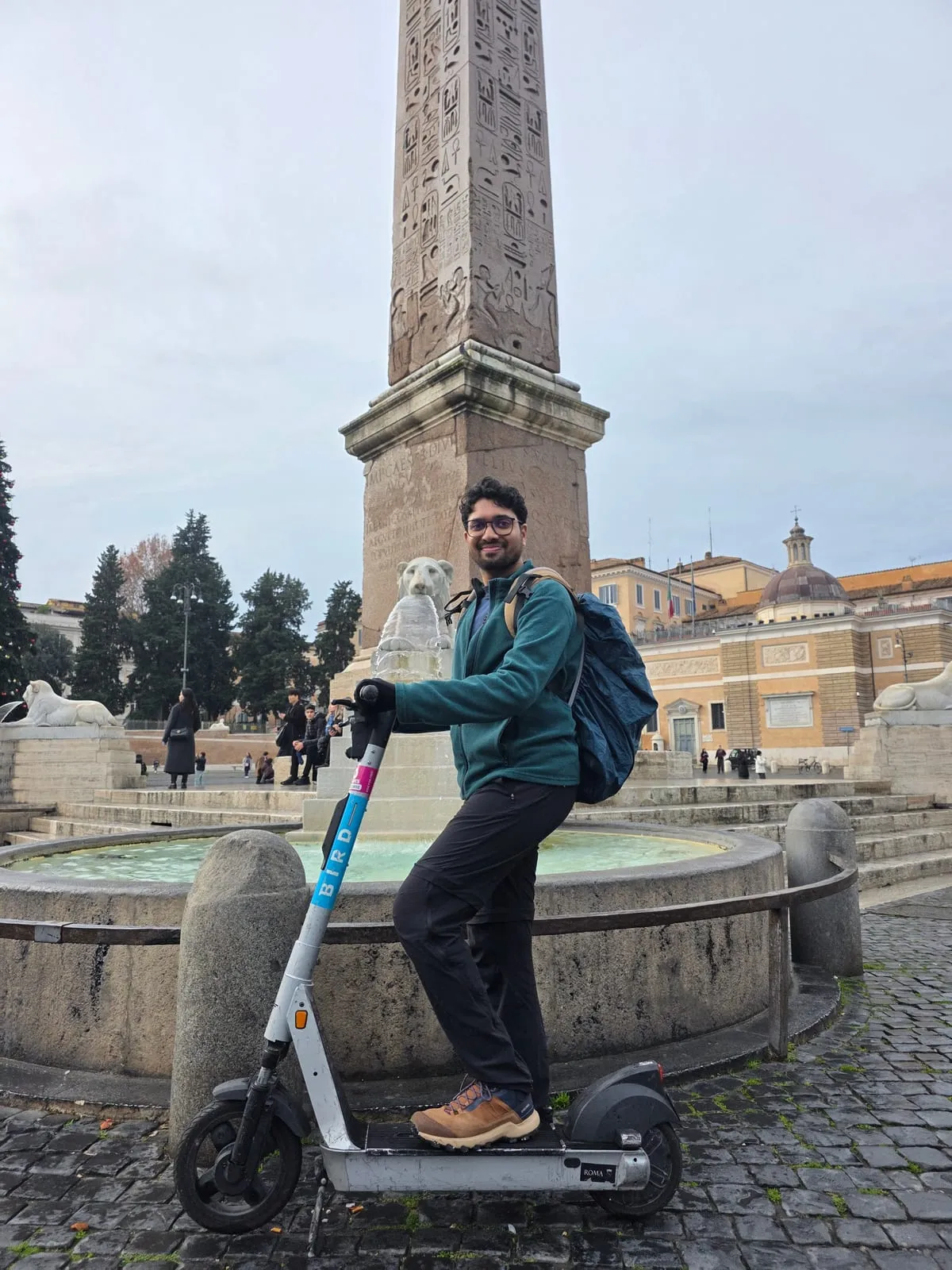 Siddhesh standing on an e-scooter in Rome's Piazza del Popolo, Italy, smiling at the camera, with the obelisk and its stone lions behind.