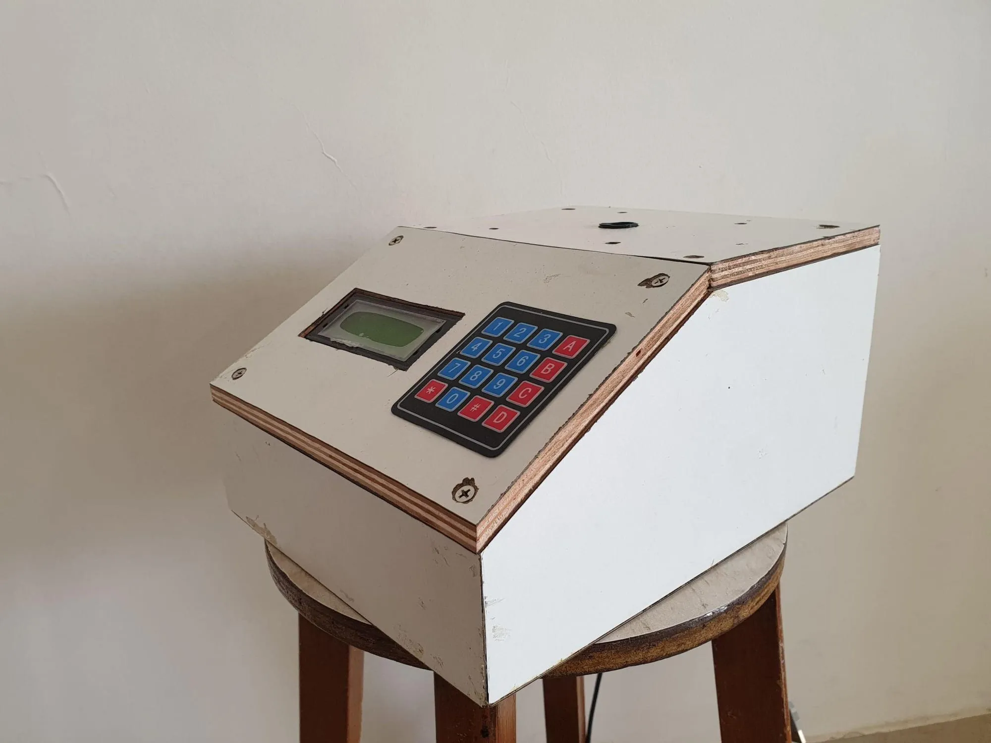 The first version of the machine, a white-painted wooden box with an LCD and keypad on its sloped front, standing on a wooden stool.